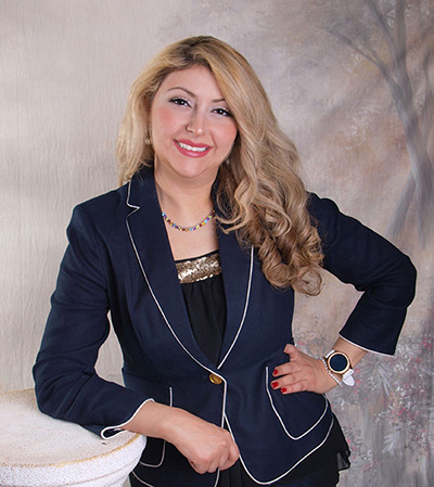 A woman with long hair wearing a blue blazer and black pants stands confidently against a backdrop with her hands on her hips.