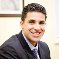 A man with a smile is wearing a suit and standing in front of an office door.