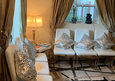 The image shows an interior space with white chairs arranged around a coffee table, adorned with glittery pillows and a silver sequin throw. A large window allows natural light into the room, which features a patterned carpet and curtains.