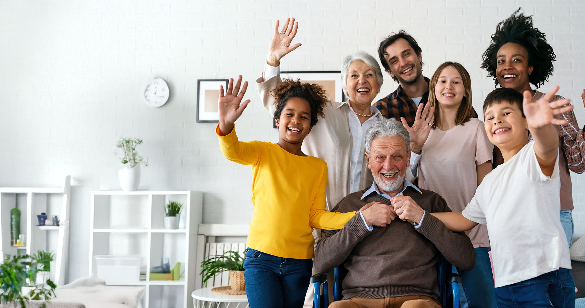 A group of people, including adults and children, waving with smiles on their faces in front of a white wall, standing around a person in a wheelchair.