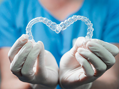 A person wearing medical gloves holds a transparent heart-shaped object, possibly representing dental implants or braces.