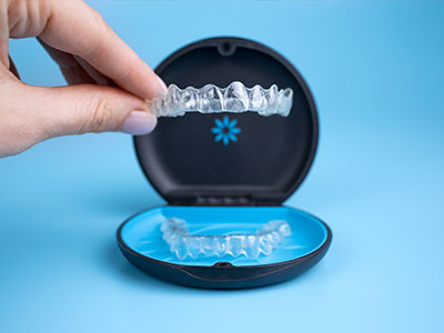 An individual s hand holding an open clear plastic case revealing transparent dental aligners inside, with a blue background highlighting the product.