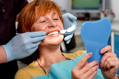 A woman sitting in a dental chair, receiving dental care with a smile on her face, while holding up a blue dental impression.