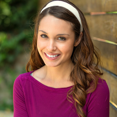 The image features a woman with long hair smiling at the camera, wearing a purple top and headband. She stands against a wooden fence.