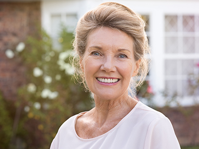 A smiling older woman with light hair, wearing makeup and a white top, stands outdoors in front of a house with a brick facade and a garden.