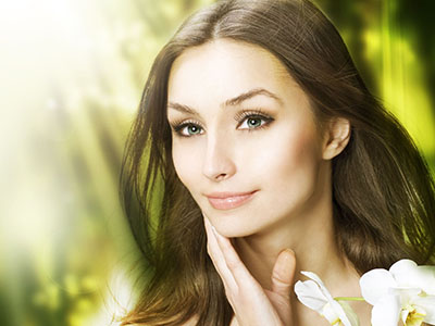 The image features a woman with fair skin, long hair, and makeup. She is looking directly at the camera with a neutral expression, against a blurred background that suggests a natural setting with greenery.