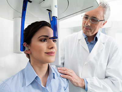 The image shows a woman seated in a dental chair with her head positioned under a device, likely for a dental procedure, while a dentist stands beside her, observing the equipment.