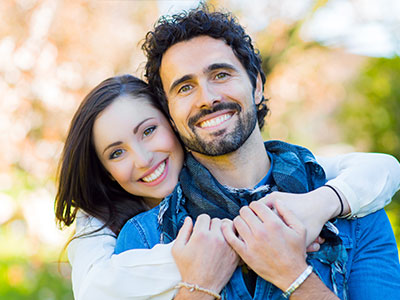 A young man and woman embrace each other warmly, smiling and appearing happy, set against a blurred outdoor background.