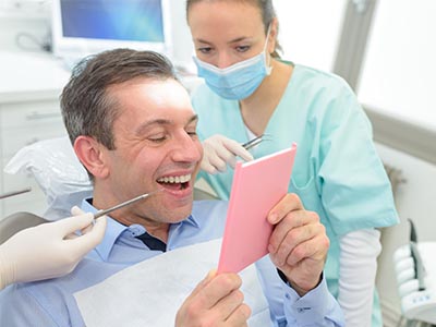 A man sitting in a dentist s chair, holding a pink card with a smile, while a dental professional looks on with a mask and surgical gloves.