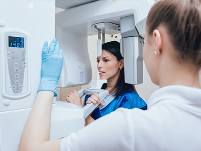 The image depicts two individuals engaged with a large medical imaging machine, possibly an MRI, with one person standing in front of the equipment and another observing from behind.