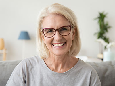 The image shows a smiling woman with short blonde hair wearing glasses, sitting on a couch in an indoor setting.