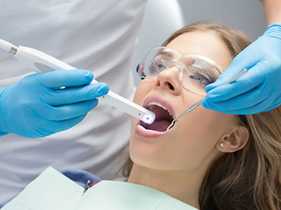 A woman receiving dental treatment with a dentist using a laser tool, while wearing protective gloves and a face mask.