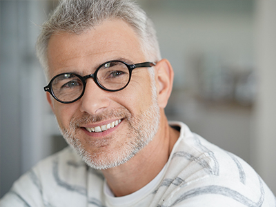 The image shows a smiling man wearing glasses, with gray hair, standing against a blurred background.