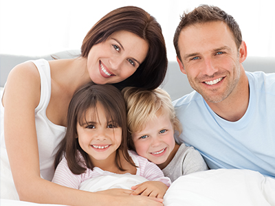 The image depicts a family with a man, woman, and three children lying together on a bed, smiling at the camera.