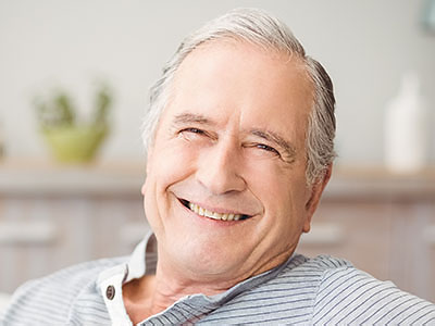 The image shows an elderly man with gray hair, smiling and looking directly at the camera. He has glasses on his head and is wearing a blue shirt. The man appears to be sitting comfortably in a chair indoors, with a warm ambiance suggested by the lighting and the presence of a plant and a vase in the background.