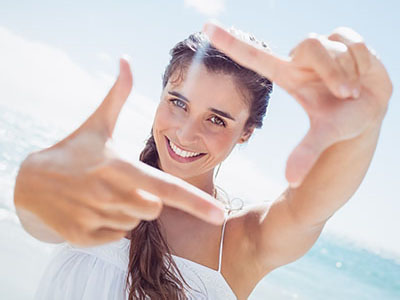 The image shows a woman with long hair smiling at the camera while holding up her hand with fingers extended, against a bright background.