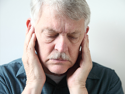 The image shows a man with his hand on his ear, possibly indicating discomfort or concern, set against a plain background.